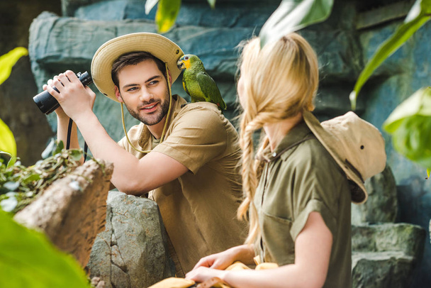 young man in safari suit with parrot on shoulder flirting with woman while hiking in jungle - Foto, Imagen