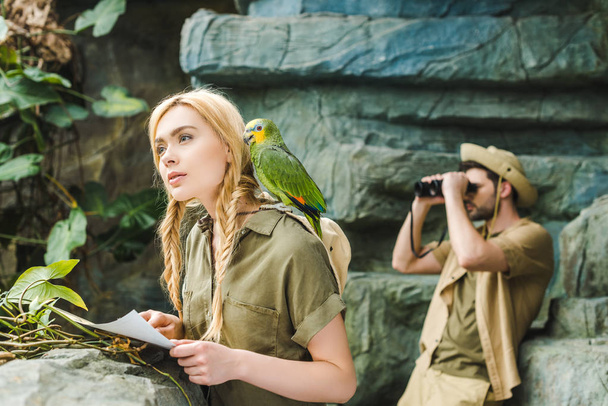beautiful young woman in safari suit with parrot and map navigating in jungle while her boyfriend looking through binoculars - Foto, afbeelding