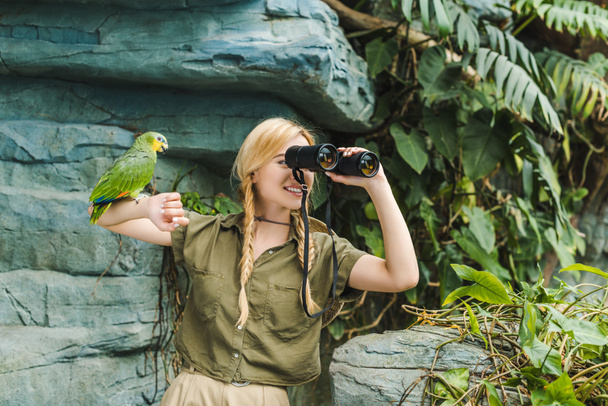 šťastná mladá žena v safari oblek s papoušek prohlížení na rameno při pohledu dalekohledem v džungli - Fotografie, Obrázek
