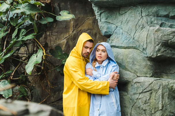 frozen young couple in raincoats embracing and trying to warm up under rocks in jungle - Foto, imagen