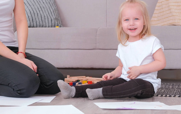 Pequeña chica rubia encantadora jugando bloques de madera con su mamá sentada cerca del sofá
. - Foto, imagen