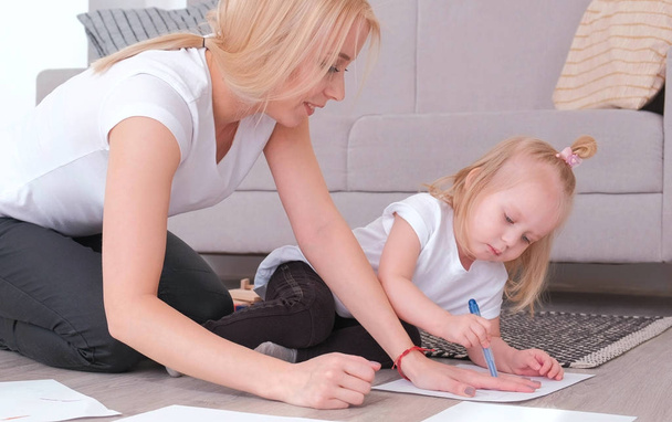 Little charming blond girl painting blue felt-pen with her mom sitting near the sofa. - Foto, afbeelding