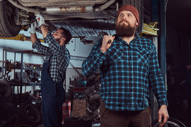 Dwóch brodatych brutalny mechanika naprawy samochodu na windę w garażu.  - Zdjęcie, obraz