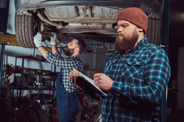 zwei bärtige brutale Mechaniker reparieren ein Auto auf einem Aufzug in der Garage.  - Foto, Bild