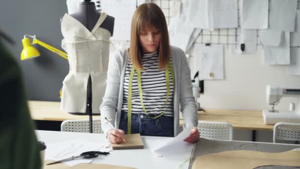 Young woman is drawing in notepad looking at clothing sketch while working in her tailoring studio. Drawings hanging on wall, bright sewing items are visible. - Кадры, видео