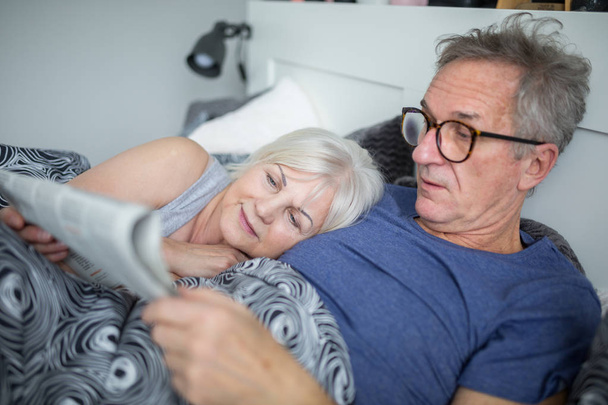 Senior man lying in bed with wife reading newspaper - Fotó, kép