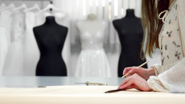 Красива дівчина модельєр малює на паперових шаблонах для вирізання тканини на манекенах, одягнених у весільні сукні. Виробник весільних суконь
. - Кадри, відео