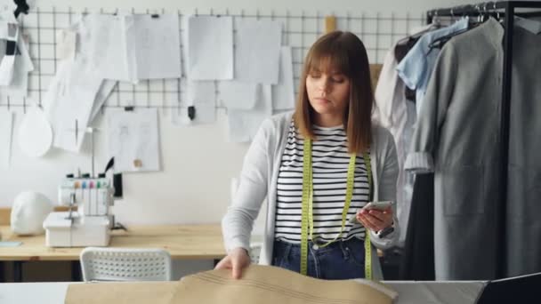 Clothing design entrepreneur is checking paper sewing drafts and looking at smartphone. She is examining every cutout carefully and thinking about future garment. - Metraje, vídeo