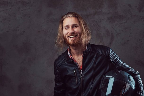 Portrait of a smiling handsome stylish redhead biker in a black leather jacket, holds motorcycle helmet, posing in a studio. Isolated on a dark background. - Photo, Image
