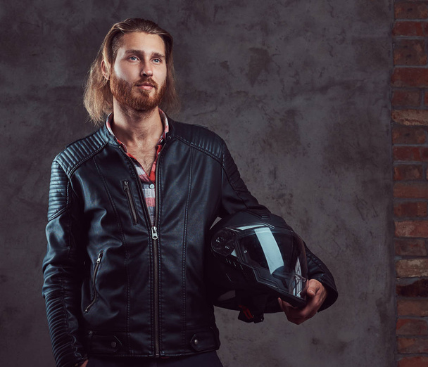 黒い革のジャケットでハンサムなスタイリッシュな赤毛バイカーの肖像画は、バイクのヘルメット、スタジオでポーズを保持しています。暗い背景に分離. - 写真・画像