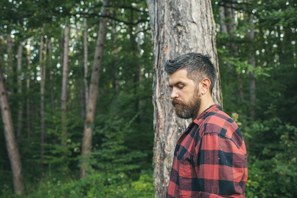 Thoughtful bearded man in lumberjack shirt alone in forest or park. Handsome hiker enjoying nature on his own. Active lifestyle, traveling and solitude concepts. - Foto, Imagem