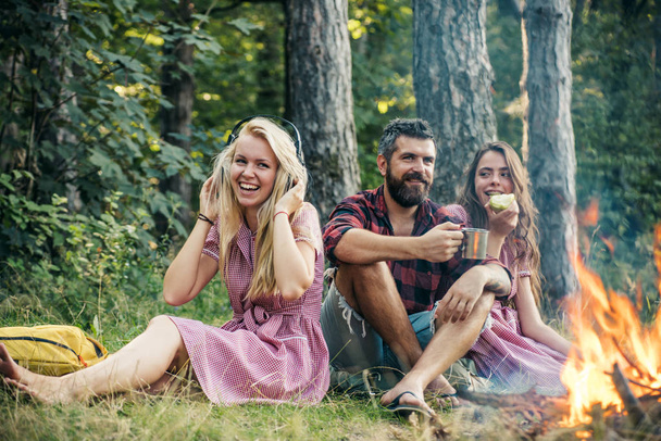 People camping at fire in forest in vintage style. Friends relax at bonfire flame with sparks. Women and bearded man at campfire. Eating food, reading book and entertainment. Summer vacation concept. - Fotografie, Obrázek