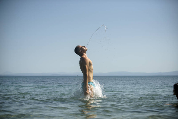Man jump out of sea with splashes of water. Vacation concept. Guy in swimwear make stream of water out of his mouth with sea water, skyline on background, rear view. Young man enjoy swimming in ocean. - Foto, imagen