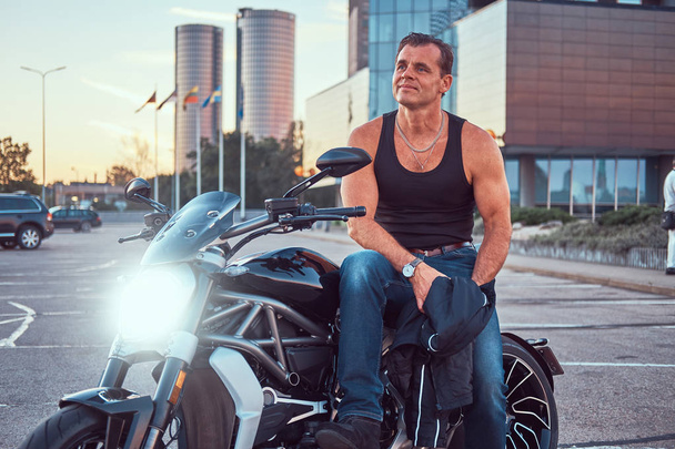 Красивий жорстокий велосипедист, одягнений в чорну футболку і джинси, сидить на спортивному мотоциклі, ностальгічний проти заходу сонця в центрі міста
. - Фото, зображення