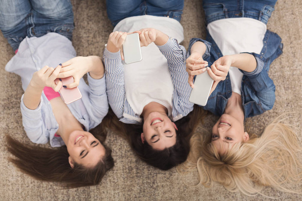 Счастливые девочки, лежащие на полу и пользующиеся смартфонами, вид сверху
 - Фото, изображение