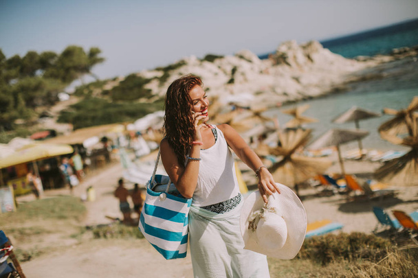 Pretty young woman with beach bag talking on mobile phone by the sea at summer - Фото, изображение