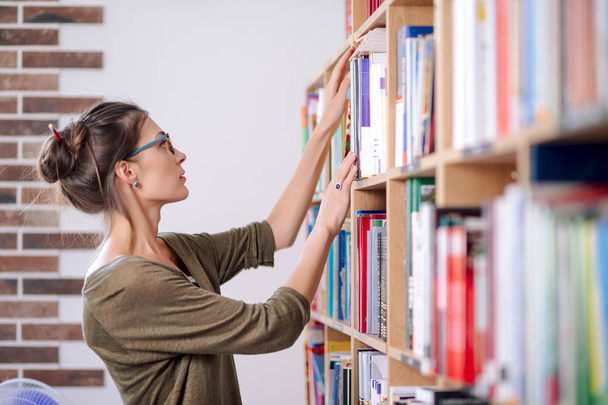 Fiatal nő visel szemüveget keres egy könyv-ra könyvespolc, én - Fotó, kép
