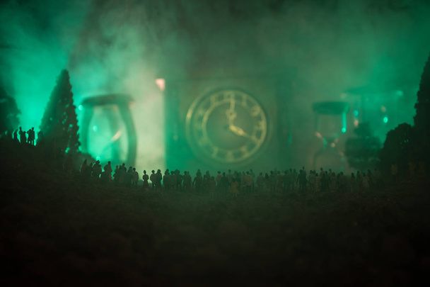 Silhouette di una grande folla di persone nella foresta di notte in piedi contro un grande orologio freccia con fasci di luce tonica su sfondo nebbioso. Concetto temporale
. - Foto, immagini