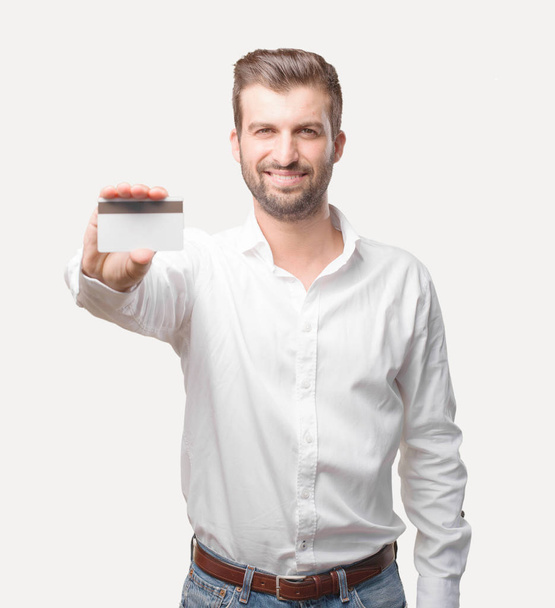Young handsome man with a credit card wearing a white t shirt, satisfied expression. Person isolated against monochrome background. - Фото, изображение