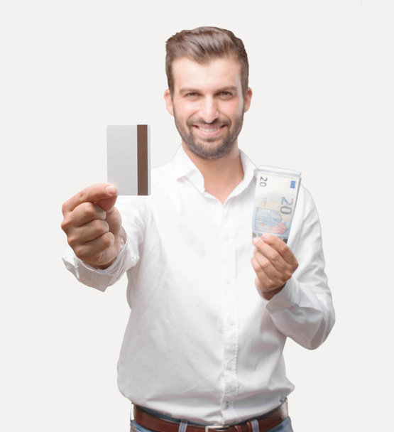 Young handsome man with a credit card and 20 euro banknotes wearing white t shirt, happy expression. Person isolated against monochrome background. - Zdjęcie, obraz