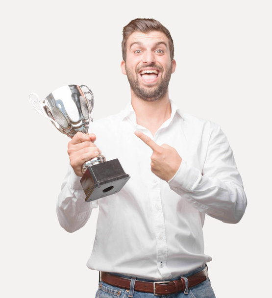 Young handsome man holding a trophy wearing white t shirt, success expression. Person isolated against monochrome background. - Фото, изображение
