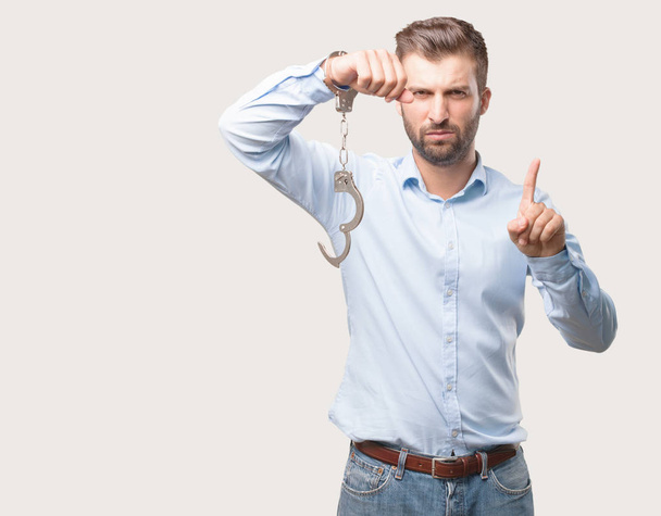 Young handsome man angry with handcuffs wearing a blue t shirt. Person isolated against monochrome background. - Φωτογραφία, εικόνα