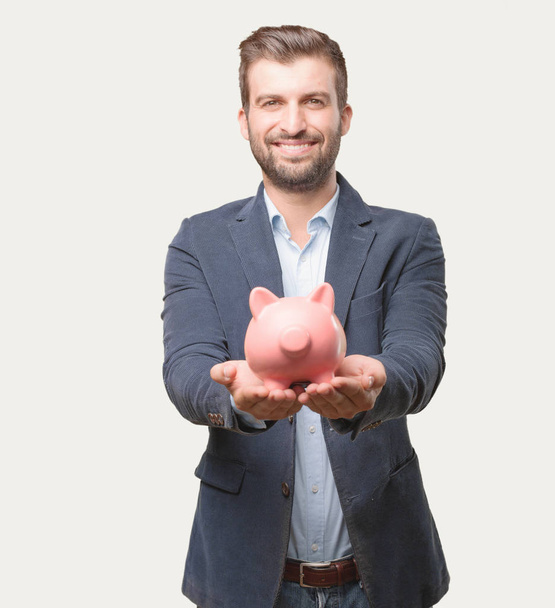 young handsome businessman, blue blazer, holding a piggy bank, happy or celebrating his success expression. Savings concept. Person isolated against monochrome background. - Фото, зображення