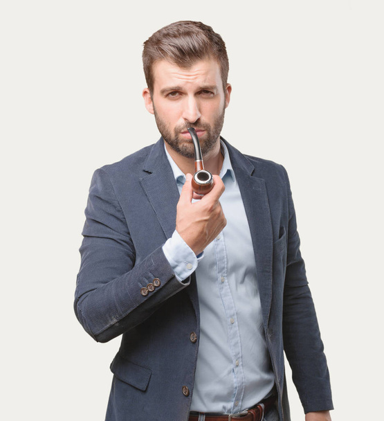 Young handsome businessman smoking with a vintage pipe, wearing blue blazer, satisfied expression. Person isolated against monochrome background. - Zdjęcie, obraz