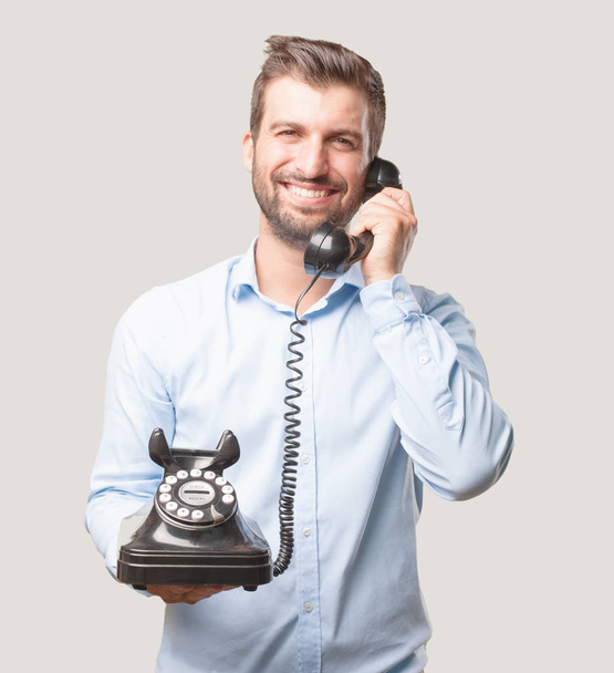 Young happy and handsome man, blue t shirt, smiling holding a vintage black telephone. Person isolated against monochrome background. - Фото, зображення