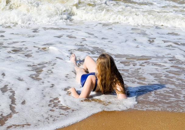 Girl in a blue bathing suit lies on the seashore and relaxes. Waves caress the girls body - Foto, Imagen