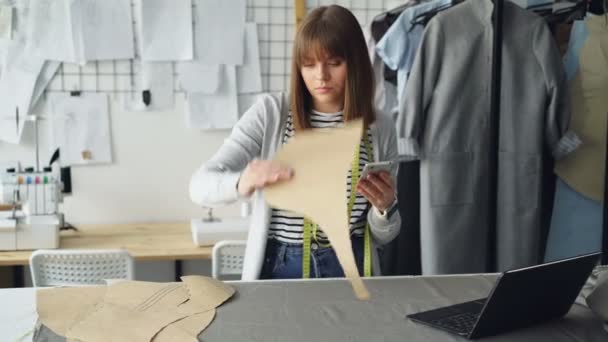 Young seamstress is checking clothing paper patterns and measuring them with tape-measure while looking at smart phone. Nice studio with garments and sewing machine in background. - Video