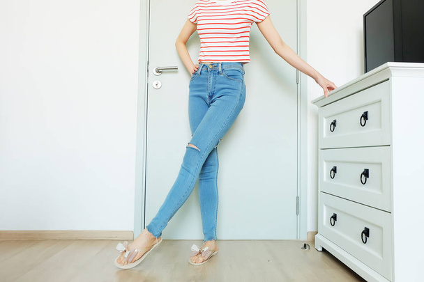 Summer Fashion Nude Bow Sandal (Footwear) and Slim Legs in The Room. Female Sexy Long Legs. Beautiful Slim Legs Woman Standing with Nude Sandals and Lack Blue Jean on Wooden Floor at Home Background - Zdjęcie, obraz