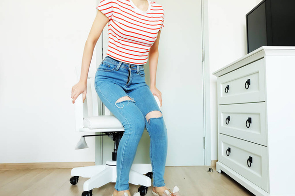 Portrait People Sit. Woman in Fashion Striped Orange Shirt and Lack Blue Jeans. Beautiful Female Sitting on White Chair at Home Background - Photo, image