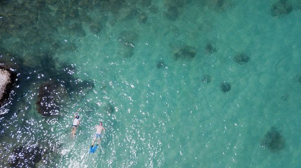 Aerea: vista aerea a persone che fanno snorkeling in onde di schiuma verde trasparente contro la linea rocciosa e fondo corallo
  - Foto, immagini