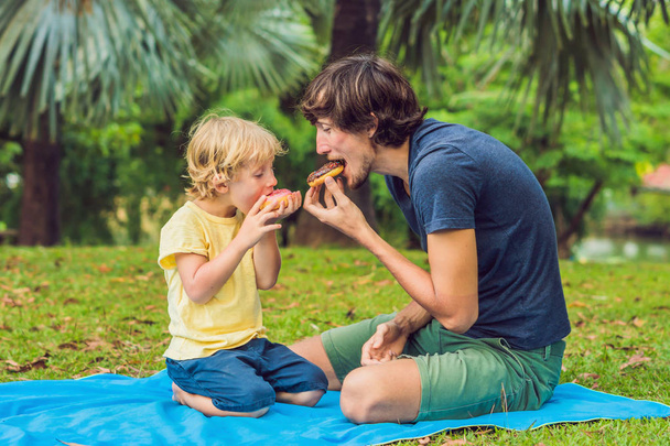 Dad and son are eating a donut in the park. Harmful nutrition in the family - Fotografie, Obrázek