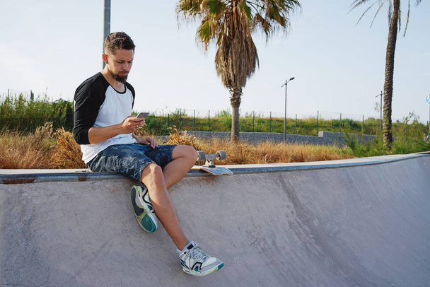 handsome young man using smartphone while having break in a skate park on a sunny summer day - Fotografie, Obrázek