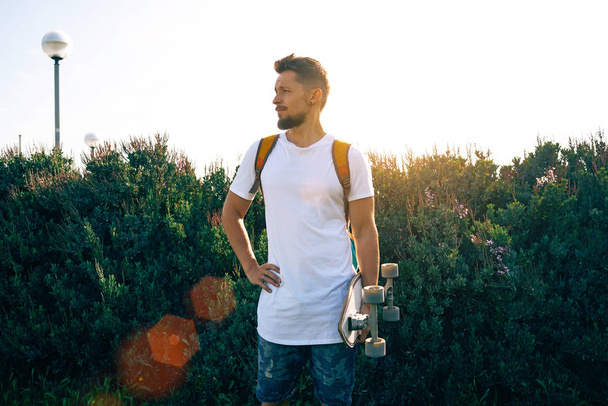 Young man with a skateboard in a blank white t-shirt standing in a bushy area - Foto, imagen