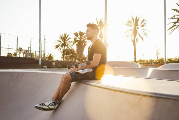 Young man in black t-shirt and shorts sitting in a skate zone with skateboard  - Fotografie, Obrázek