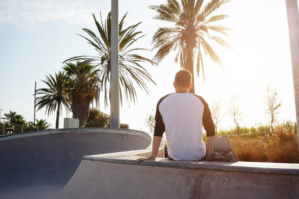 A male in a blank white long sleeve t-shirt is sitting with the back to the camera. Young man with the skateboard is sitting in the urban zone while relaxing on a sunny background - Фото, зображення