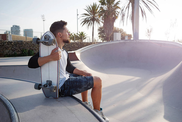 Young bearded man in a street wear is sitting with the skateboard in the urban zone. A skater is having rest in the urban zone on a sunset - Fotografie, Obrázek