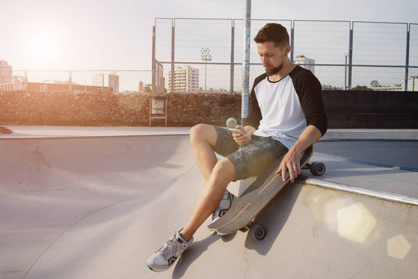 Young man is holding a mobile phone while sitting in a skate zone. A guy is surfing the web by the smartphone while sitting on a sunset background - Fotoğraf, Görsel