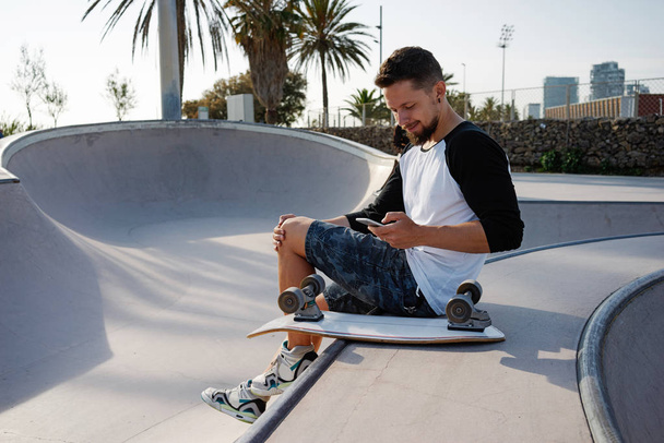 Young man is holding a mobile phone while sitting in a skate zone. A guy is surfing the web by the smartphone while sitting on a sunset background - Foto, immagini