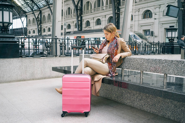 Beautiful Caucasian woman with luggage using smartphone while sitting in train station interior - Фото, изображение