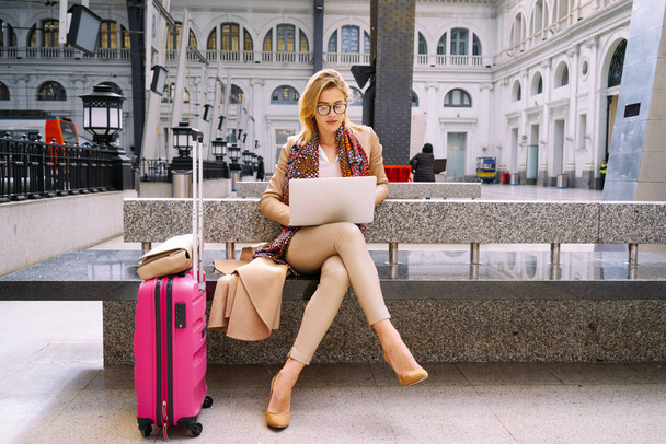 Beautiful young woman using laptop while sitting with luggage in train station - Photo, image