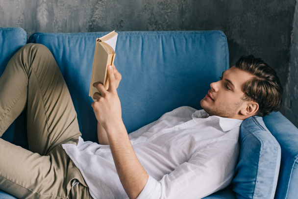 Junger Mann liest Buch, während er auf dem Sofa liegt - Foto, Bild