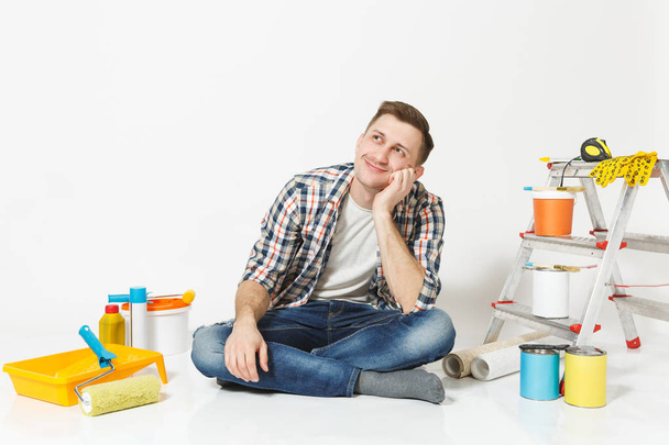 Man in casual clothes dreamily looks up, sitting on floor with instruments for renovation apartment isolated on white background. Wallpaper, gluing accessories, painting tools. Repair home concept. - Photo, Image
