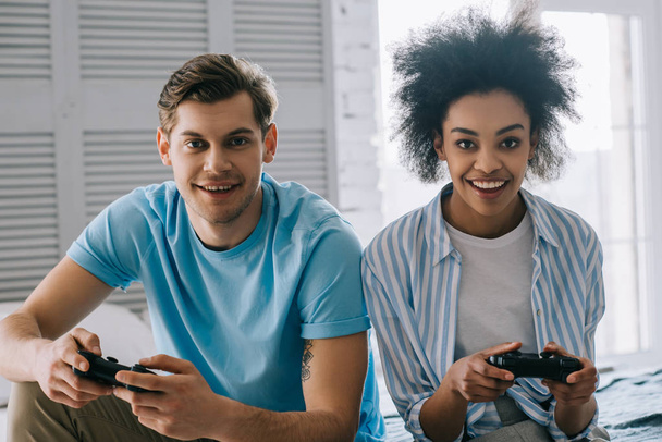 Multiracial couple with joysticks playing video game while sitting on bed - Foto, Imagem