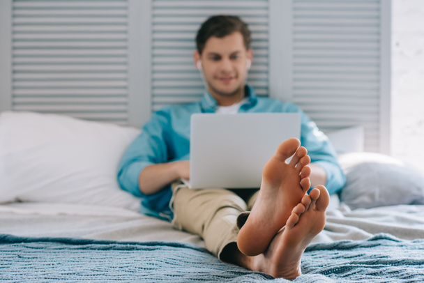 Nahaufnahme der Füße des Mannes mit Laptop, während er zu Hause auf dem Bett liegt - Foto, Bild