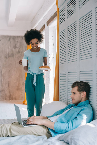 African american girl standing with breakfast by her boyfriend in bed with laptop - Foto, afbeelding