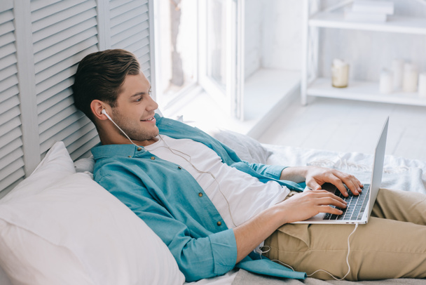 Smiling man in earbuds using laptop while lying on bed at home - Photo, Image